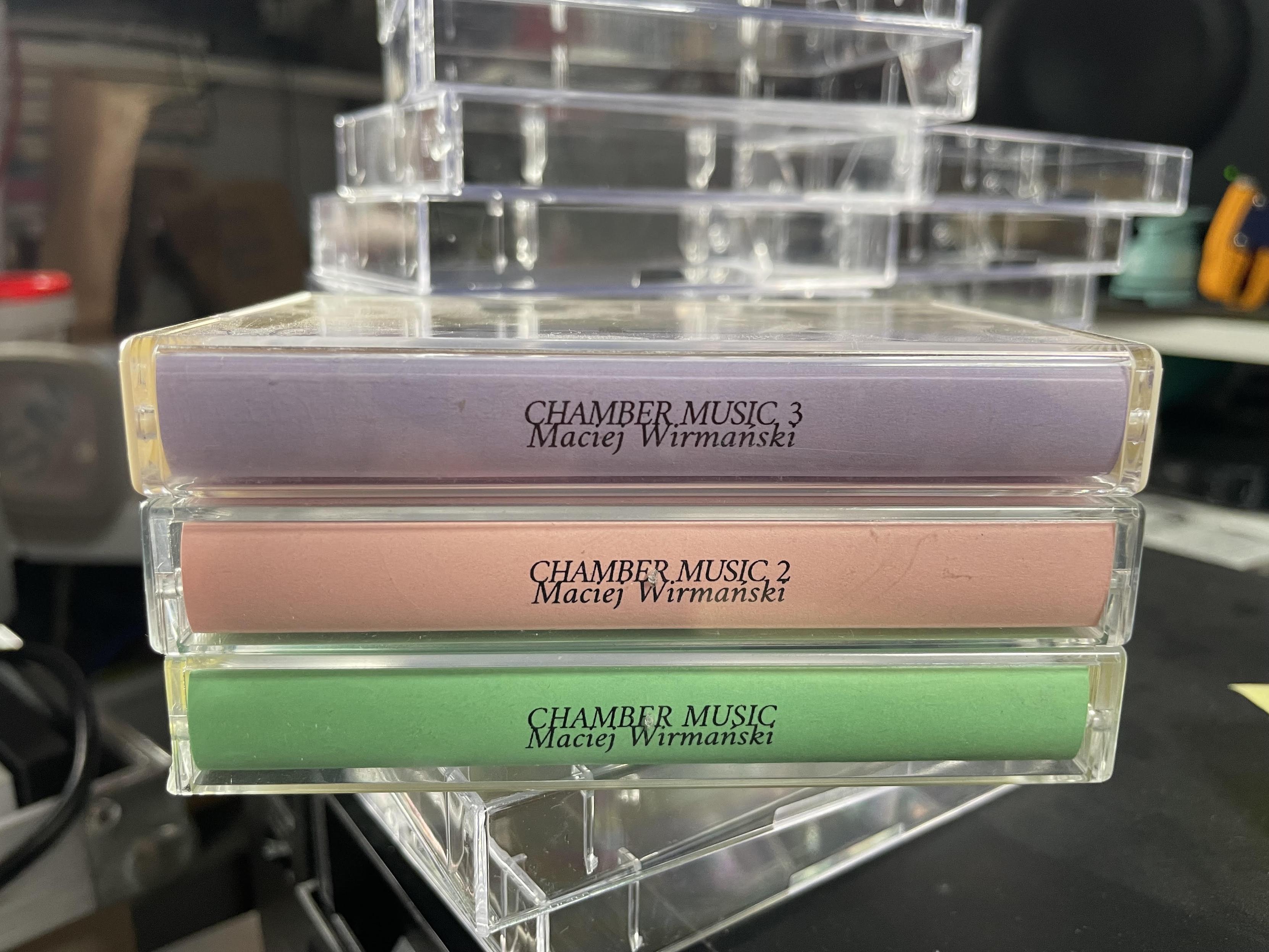 stosik trzech kaset obróconych brzegiem w stronę kamery. kasety są różowe, fioletowe i zielone, i napisane na nich jest "Maciej Wirmański, Chamber Music, Chamber Music 2, Chamber Music 3", z tyłu większy stos przezroczystych pudełek na kasety w nieładzie