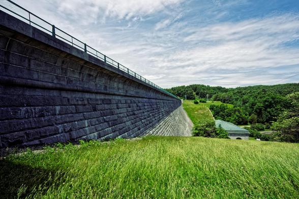 On the left a great dam made of large granite blocks extends from the front to the back with a long grass lawn immediately in front of us and field, water and forest across a great gulf with high altitude clouds high overhead