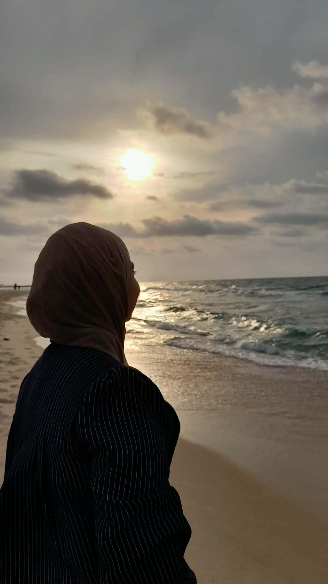 Naeema in hijab facing away from the camera, standing on a beach and looking at the ocean and sunset.