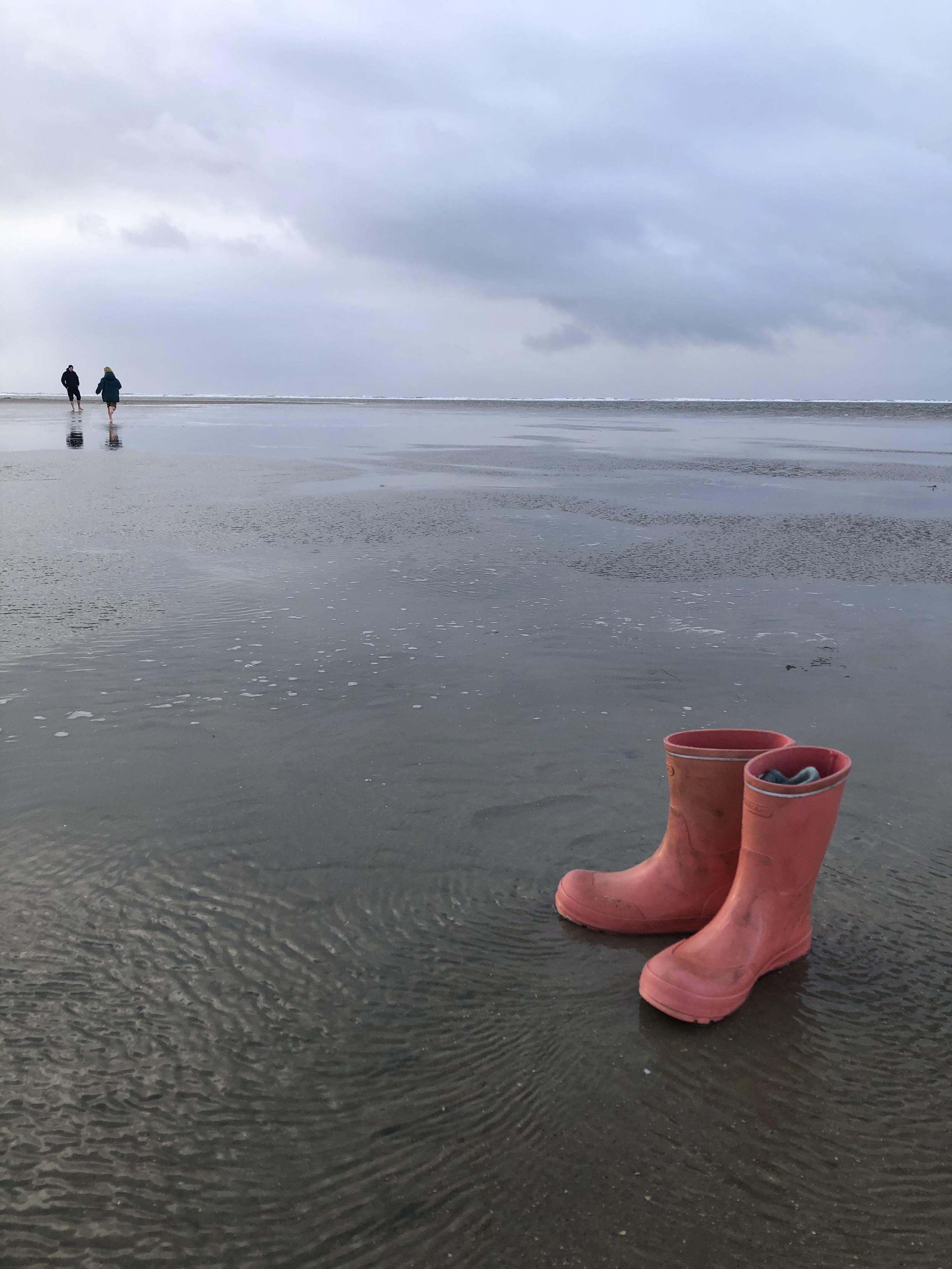 Ein weitläufiger, nasser Sandstrand unter einem bewölkten, grauen Himmel. Im rechten Vordergrund stehen ein Paar rosa Gummistiefel aufrecht im feuchten Sand mit Wellenstrukturen. In einem der Stiefel ist ein kleiner blauer Gegenstand sichtbar. Die nasse Oberfläche des Strandes spiegelt den Himmel wider. Auf der linken Seite im Hintergrund laufen zwei Personen in die Ferne über das flache Gelände.