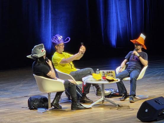 Seated on stage: Pablo in a baby shark hat, Guido in a octopus hat, and Łukasz in a. VLC traffic cone hat. 