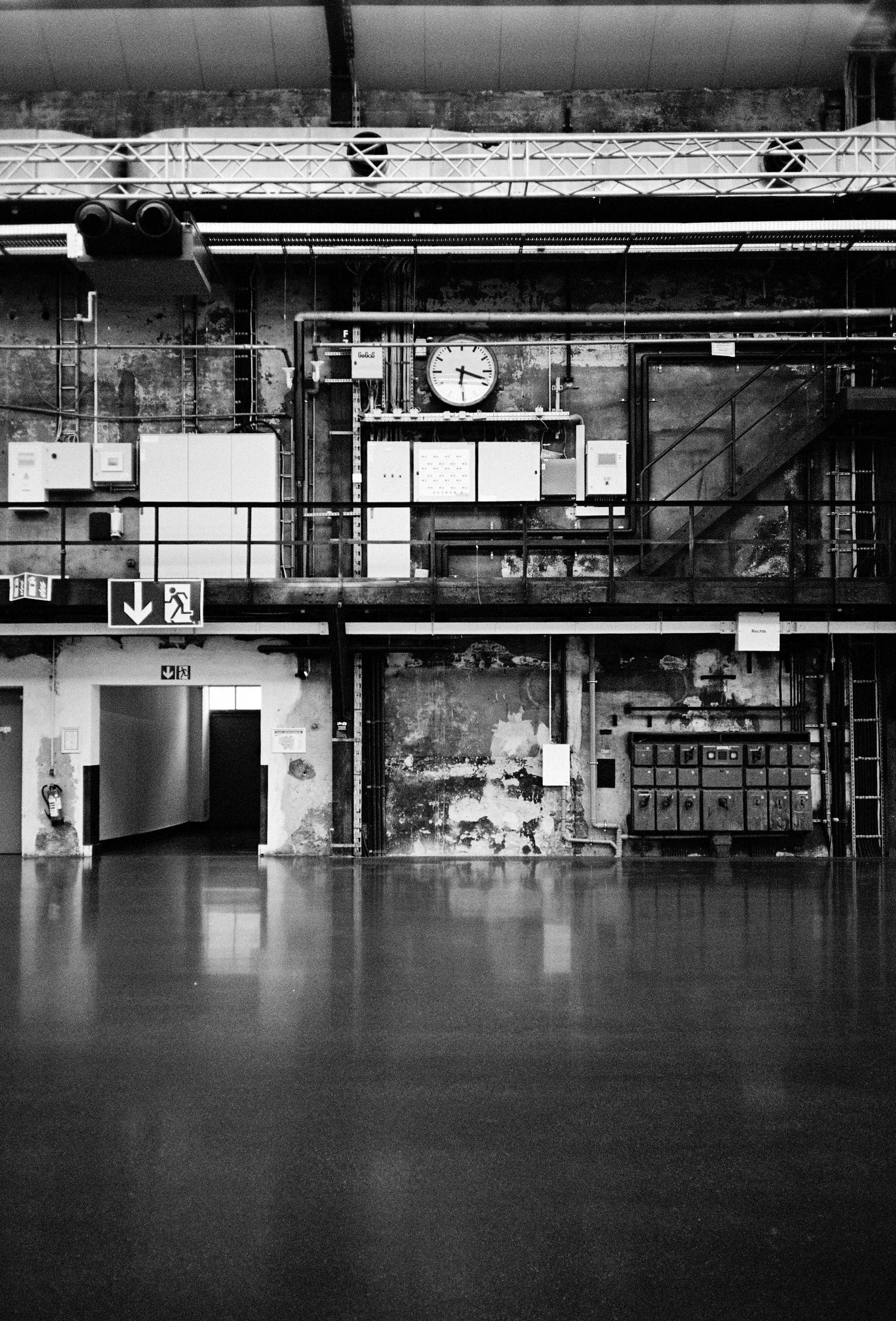 Blick auf eine interessante rustikale Industriewand, in deren Zentrum eine analoge Uhr hägt. Die Wand hat Durchgänge, Treppen, Kästen zu bieten