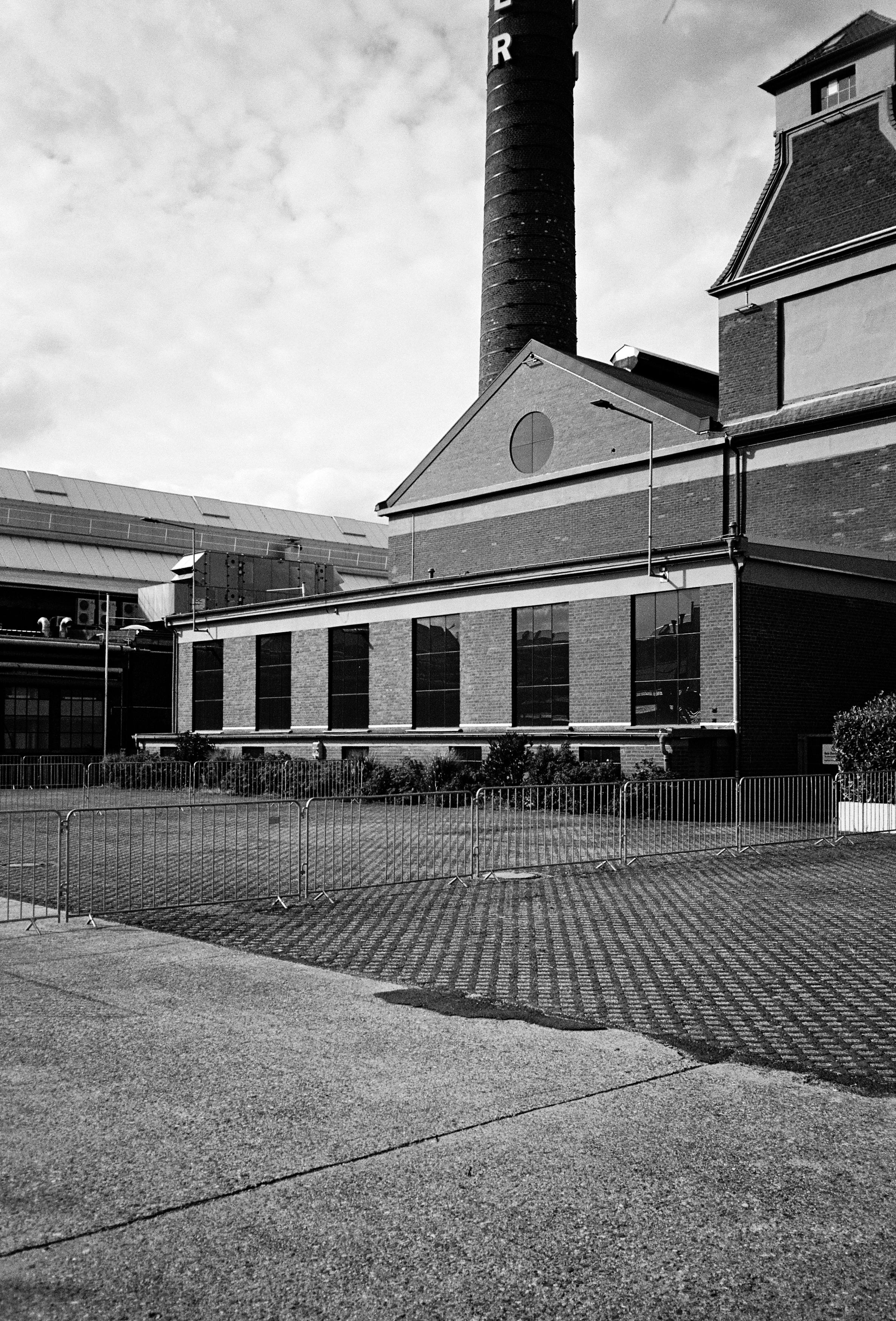 Außenansicht eines Industriegebäudes, von schrägrechts aufgenommen. Im Hintergrund ein Schornstein. Vor dem Gebäude eine mit Metallgattern abgesperrte, gepflasterte Freifläche