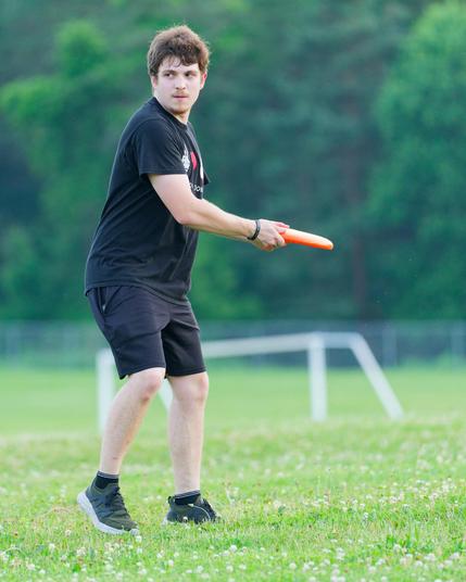 Man with black shirt holds orange frisbee with his weight mostly on one foot with his eye locked on a target in a clover-filled short-cropped lawn with a white soccer goal in the far background, a chain link fence, and a wall of lighter and darker trees behind