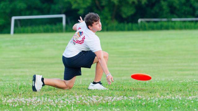 Young man with black shorts and white shirt is reaching down near an orange frisbee floating close to the ground with a band of clover just in front of him and signs of a soccer facility at the edge of the forest behind him