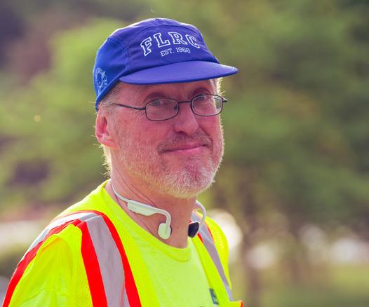 Man with white hair and beard,  blue billed cap with white outlined FLRC lettering,  around-the-neck headphones and a high-viz vest with blurry trees behind