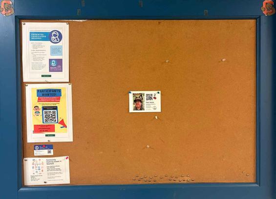 Corkboard covered surrounded by blue-painted wood has a row of mostly letter-size posters on the far left and a neat row of tacks on the bottom. Exactly in the center find a 4x6 card with a picture of a man wearing a boonie cap with fox ears on the left and a QR code business card on the right. Really!