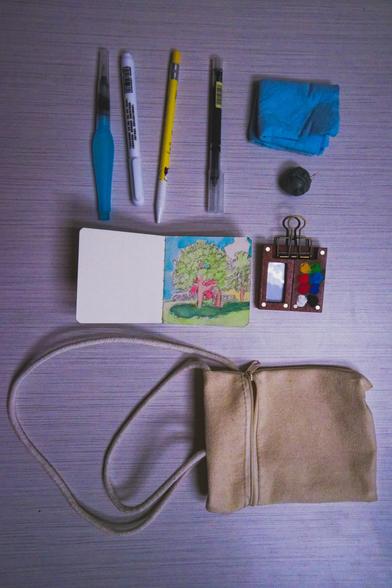 A kit for doing watercolors on the go that includes a watercolor brush with a watercolor resivoir, a white pen, a thick colored pencil, some other pen, cleaning cloth, kneaded eraser, little book with a simple watercolor of a tree I did, and a carrying bag on a table with fine lines and surprisingly moody shadows