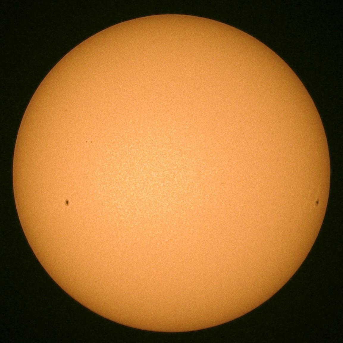 A bright orange sun against a dark background, with two small dark spots visible on its surface indicative of sunspots.