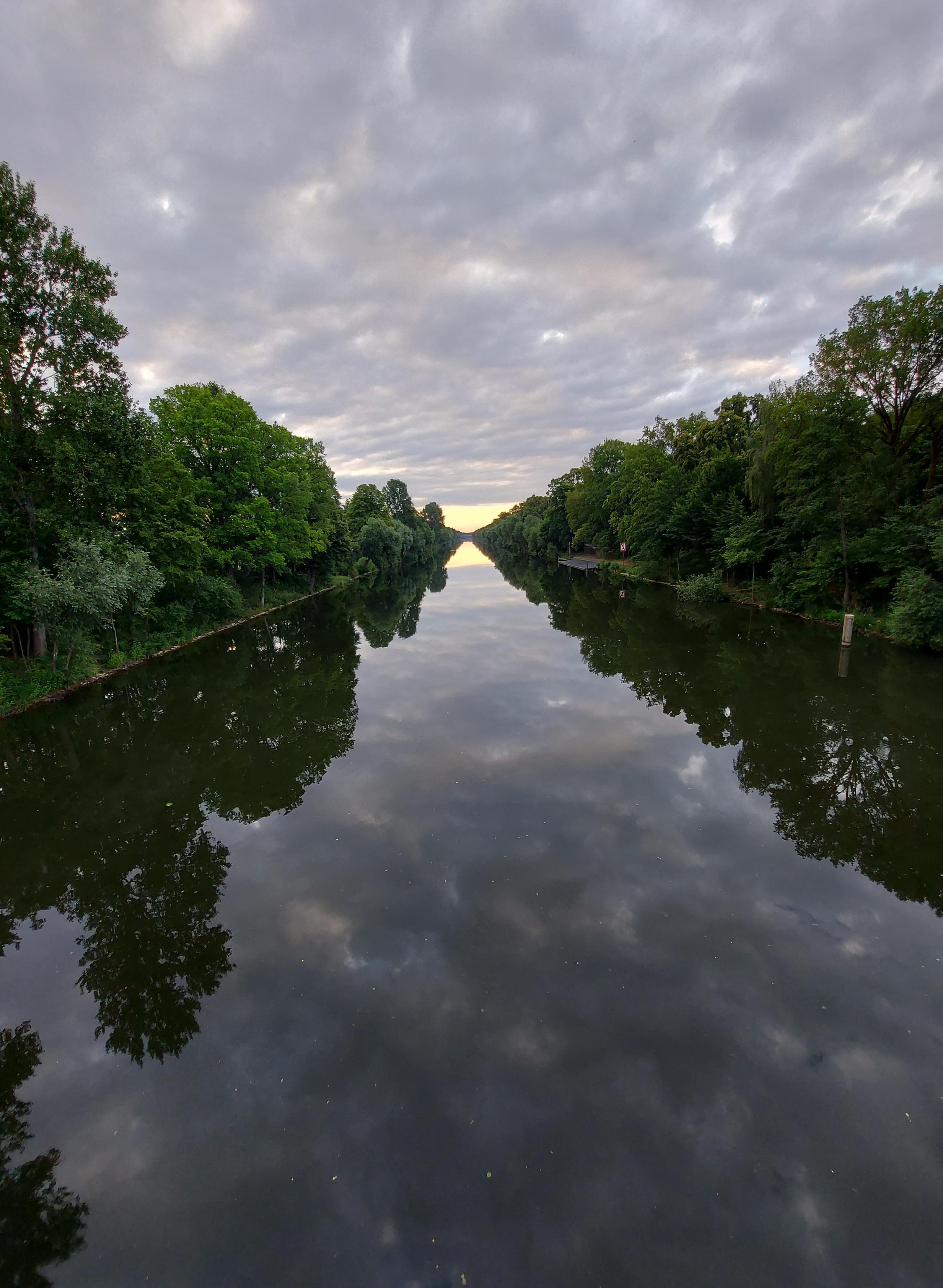 Ein stiller baumgesäumter Kanal am frühen Morgen. Blick nach Osten. Der grau-wolkige Himmel spiegelt sich, er wirkt schwer.