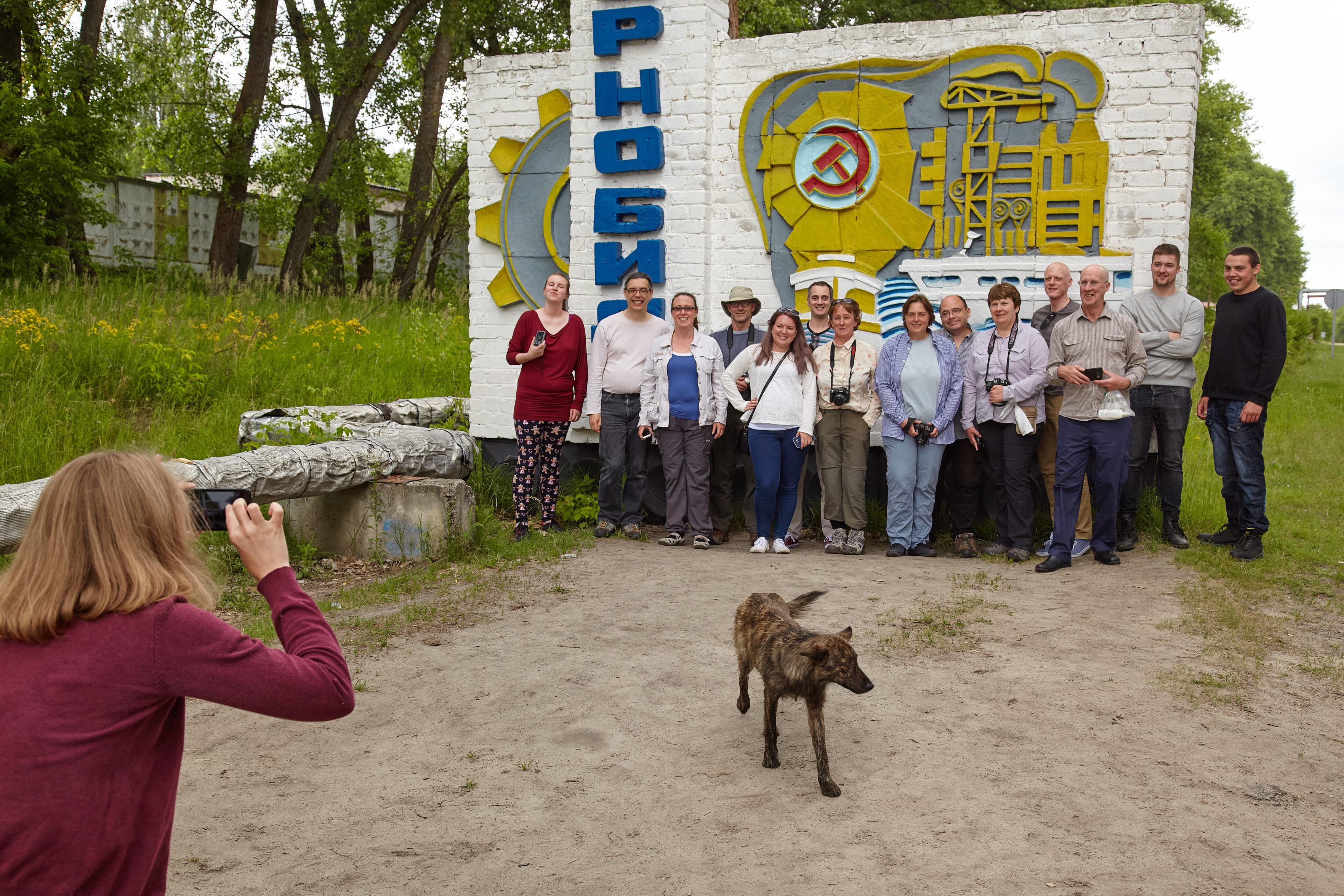 A tour group poses for a photo at Chornobyl's town sign while a stray dog trots into the frame between them and the photographer.