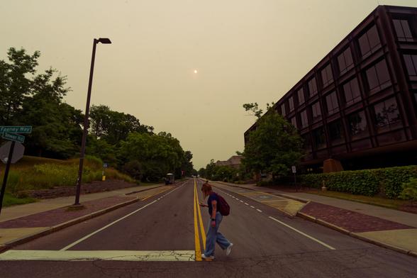 Woman with obscured face looking at cell phone crosses Tower road in front of Uris Hall under ominous strange sky with the sun just a tiny point in the sky