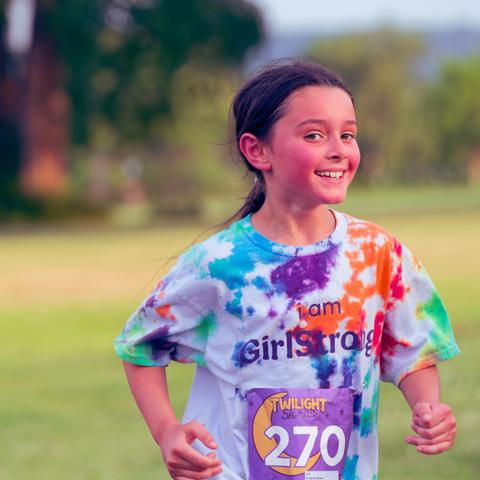 Girl with dark hair tied back is smiling and has tie-die shirt that reads "I am Girlstrong" with big smile making eye contact with me with short cropped grass and a row of trees before a view across the lake of a distant hill.