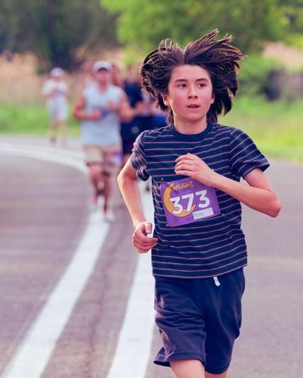 Runner with long hair flying up with dark blue shirt with fine horizontal lines followed by many more runners with various shades of green plants in the distance