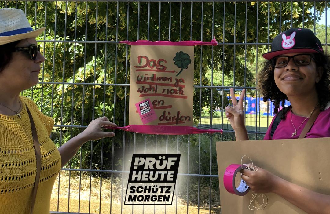 Zwei Prüfende haben gerade ein Plakat aufgehängt "Das wird man ja wohl noch PRÜFen dürfen" 
Darunter ist das Logo von PRÜF
"PRÜF
HEUTE
SCHÜTZ
MORGEN"
Das "P" von PRÜF besteht aus einem Ausrufezeichen und einem passenden Bogen am oberen Ende.