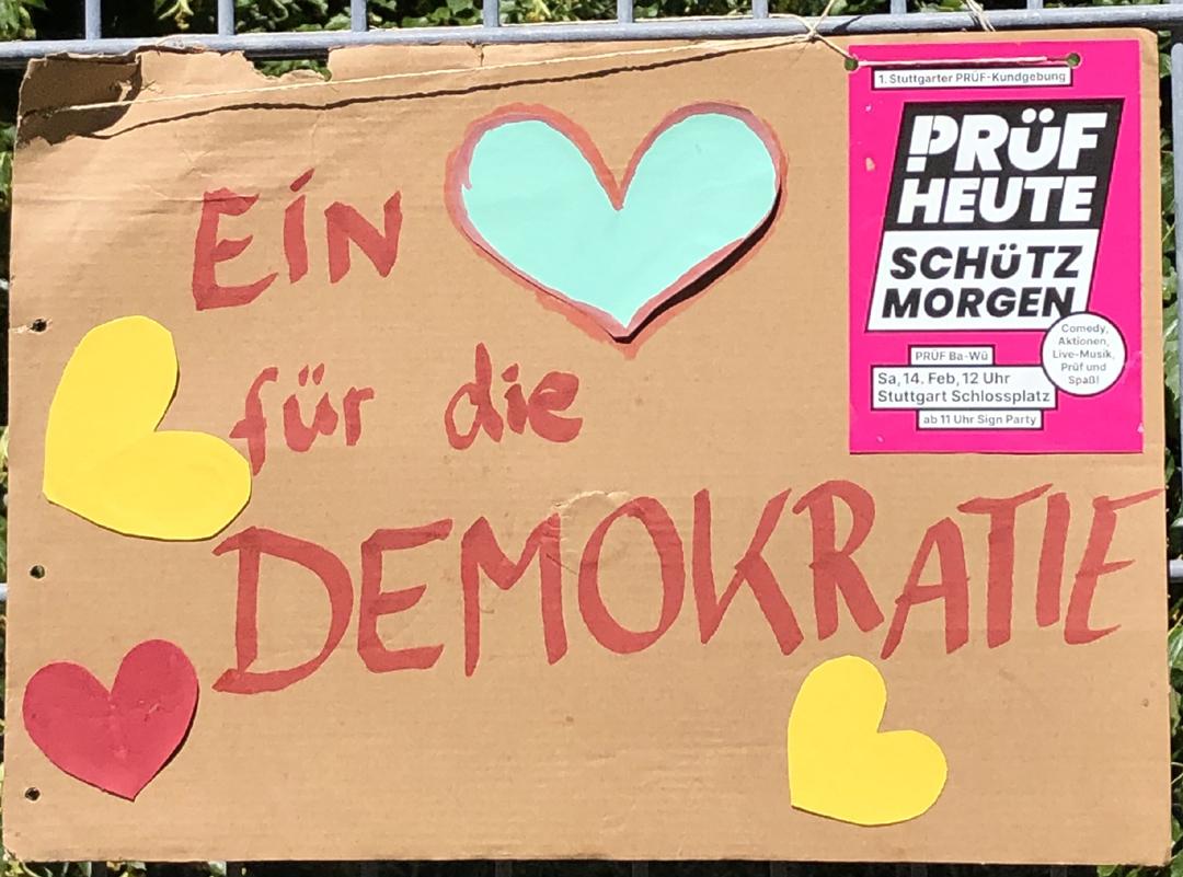 Ein Plakat mit vier bunten Herzen "Ein Herz für die DEMOKRATIE"
Oben rechts ein PRÜF-Sticker mit dem Logo von PRÜF
"PRÜF
HEUTE
SCHÜTZ
MORGEN"
Das "P" von PRÜF besteht aus einem Ausrufezeichen und einem passenden Bogen am oberen Ende.
