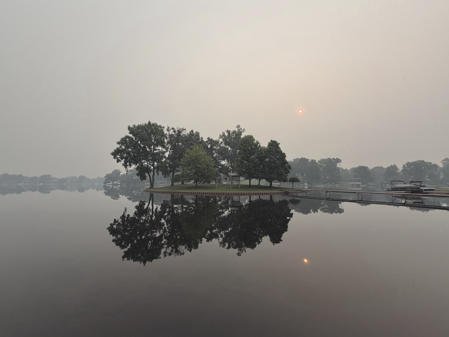 Photograph of a small island in a reflective lake a couple of hours after sunrise with a thickly smoky sky behind and the sun behind it still slightly obscured by the smoke and reflected in the lake.