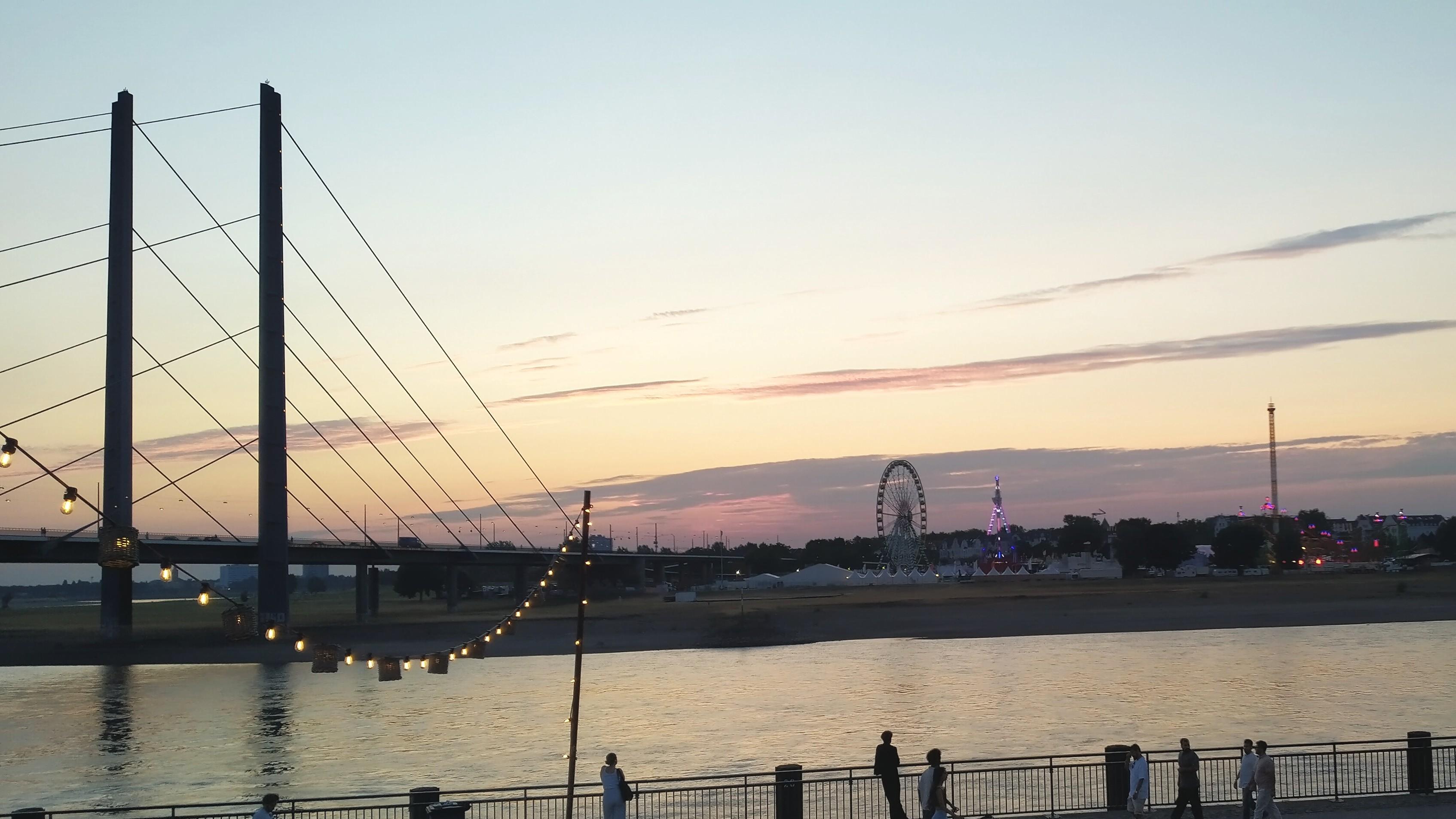 Foto des Rheins bei Düsseldorf bei niedrigem Wasserstand in abendlichem Licht unter fast wolkenlosem, dämmrigem Himmel. Links die Kniebrücke und im Hintergrund die Rheinwiesen von Oberkassel mit der Rheinkirmes.