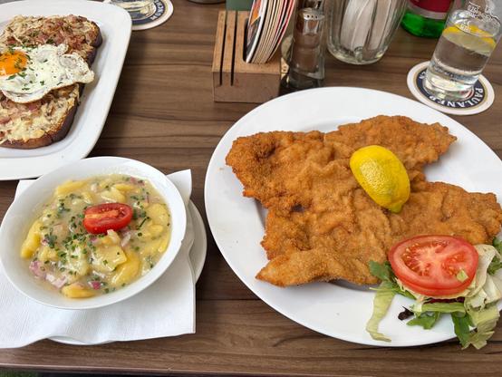 Plates set on a restaurant table. A big white plate on the right has a large piece of flat, breaded meat (Wiener schnitzel) with a slice of lemon on top. In the lower left is a bowl of potato salad with a red tomato on top