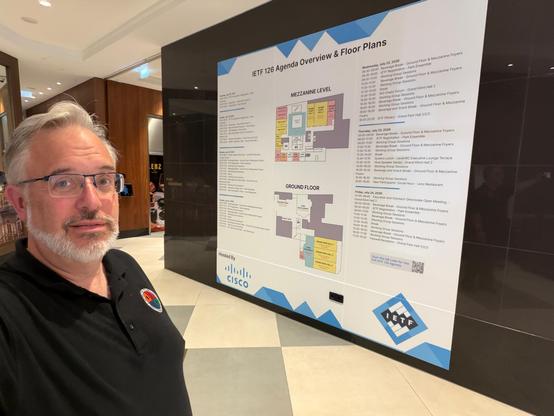 A photo of Dan York on the left and a large black wall on the right with a large white sign showing the hotel floor plan in the middle and an overview of the IETF 126 agenda in columns on the left and right. The sign stretches for most of the wall and from near the ceiling to down near the floor. 