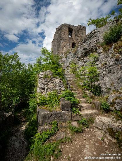 This image captures a dramatic and historic scene featuring the ruins of a stone tower, likely part of an old castle or fortress, perched on a rocky cliff. The tower, constructed from weathered gray stone, stands tall against the backdrop of a bright blue sky dotted with fluffy white clouds. The structure has an arched window, hinting at its ancient architectural style and the passage of time.
A narrow, winding stone staircase with a metal handrail leads up to the tower, inviting the viewer to …