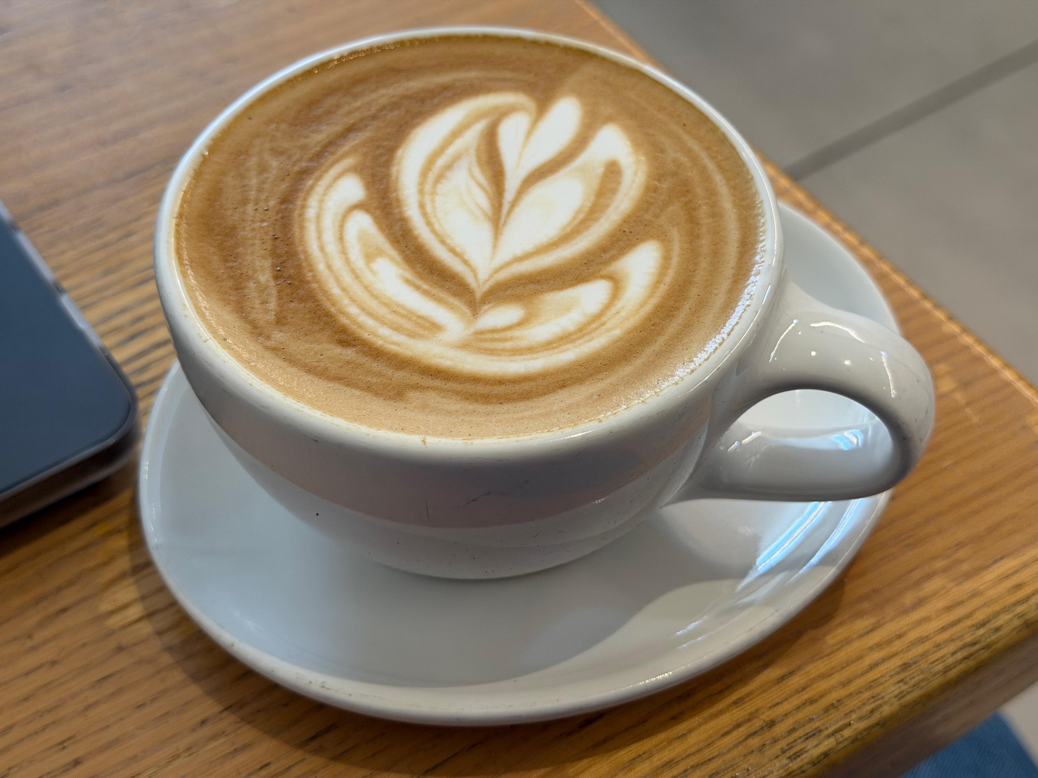 A picture of a latte in a ceramic cup on the corner of a wooden table with detailed latte art resembling an abstract heart. The corner of a laptop can be seen on the table next to it next to it. The floor in the background is blurry.