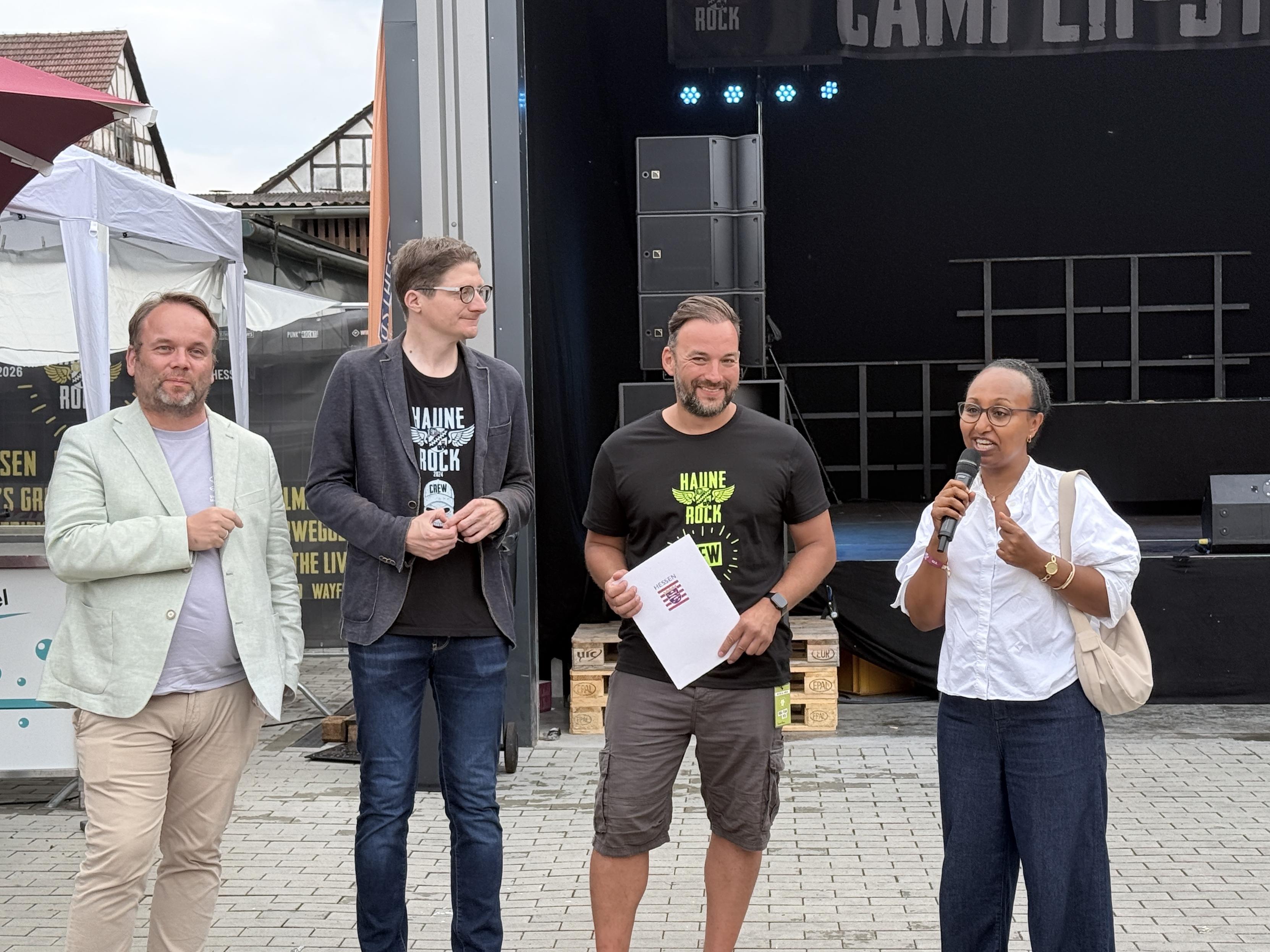 Eine Gruppe von vier Personen steht bei einer Veranstaltung im Freien vor der Bühne. Eine Person (Awet) spricht in ein Mikrofon, während die anderen zuschauen. Der Schauplatz scheint ein Festival oder ein Konzert zu sein, mit einer Bühne und Ausrüstung, die im Hintergrund sichtbar sind. Es sind Landeskulturminister Gremmels, der Bürgermeister von Haunetal Timo Lübeck, der Geschäftsführer von Haunerock und Awet Tesfaiesus.