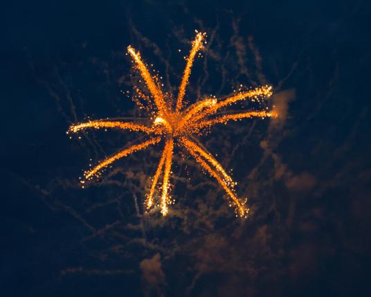 Firework with streamers yellow at the base turning white to the tips that are thick and sparkly with illuminated irregular smoke trails behind
