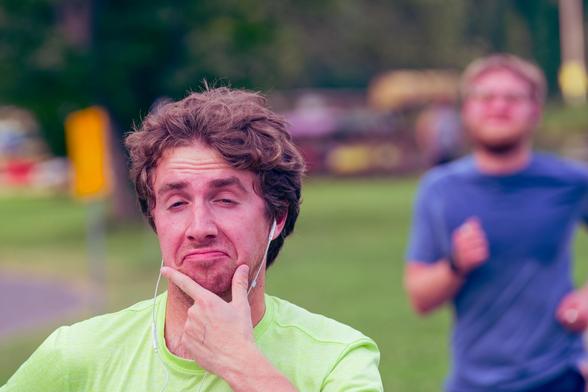 Man wearing earbuds and yellow shirt makes a funny expressing with his hand on his chin while another blurred out runner in a blue shirt runs up from behind