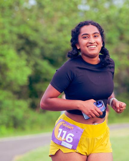 Smiling woman has black top and yellow shorts with bib number 117 and an open midriff with twinbraided black hair with light colored trees behind