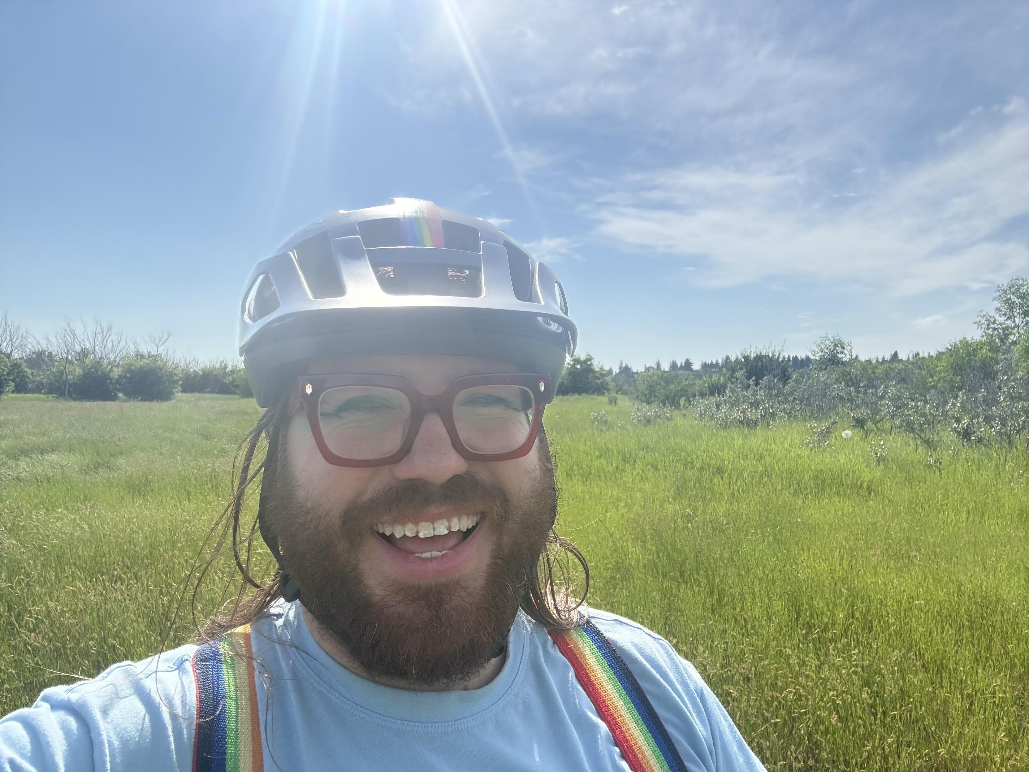 Selfie of a smiling fellow in a bike helmet and rainbow suspenders. The sun shines behind, washing out the photo a bit.