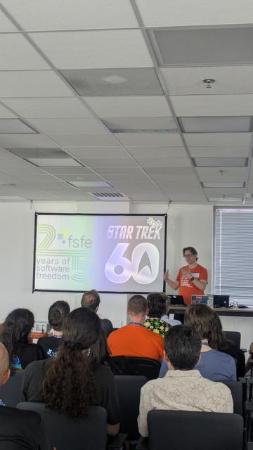 A white male talking to a sitting audience. There is a screen with a slide with the fsfe 25 years logo and star trek 60