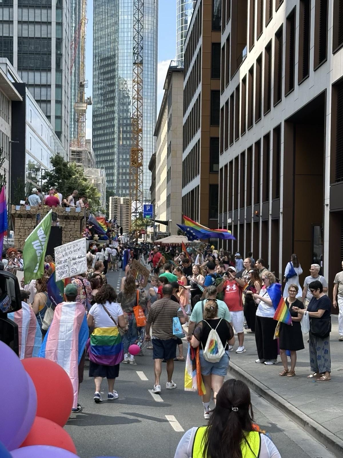 Eine lebhafte Feier mit einer großen Menge, die an einer Pride-Parade teilnimmt. Menschen halten verschiedene Regenbogenfahnen und Schilder in der Hand, während sie eine Stadtstraße entlanggehen, die von modernen Gebäuden flankiert wird. Ballons sind im Vordergrund sichtbar, was zur festlichen Atmosphäre beiträgt.