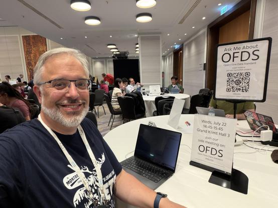 A smiling person with glasses wears a dark t-shirt and is seated at a table in a conference setting. Laptops are on the table, and a sign promotes a side meeting about the Open Fibre Data Standard (OFDS) in Grand Klimt Hall