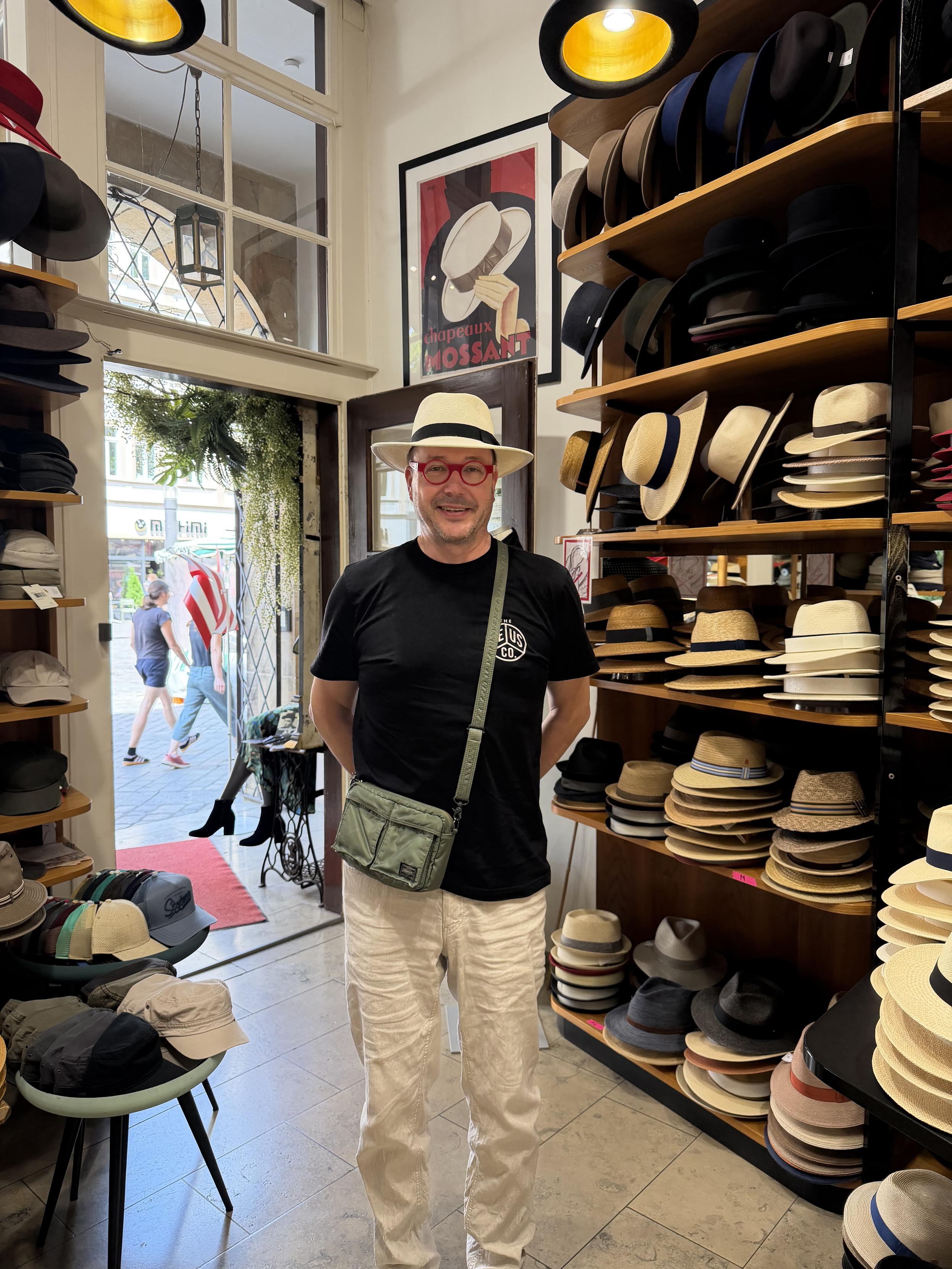 Ein Mann im Hutgeschäft mit Sonnenhut, schwarze T-Shirt, beige Hose, rote Brille, lächelnd vor Hutregal.