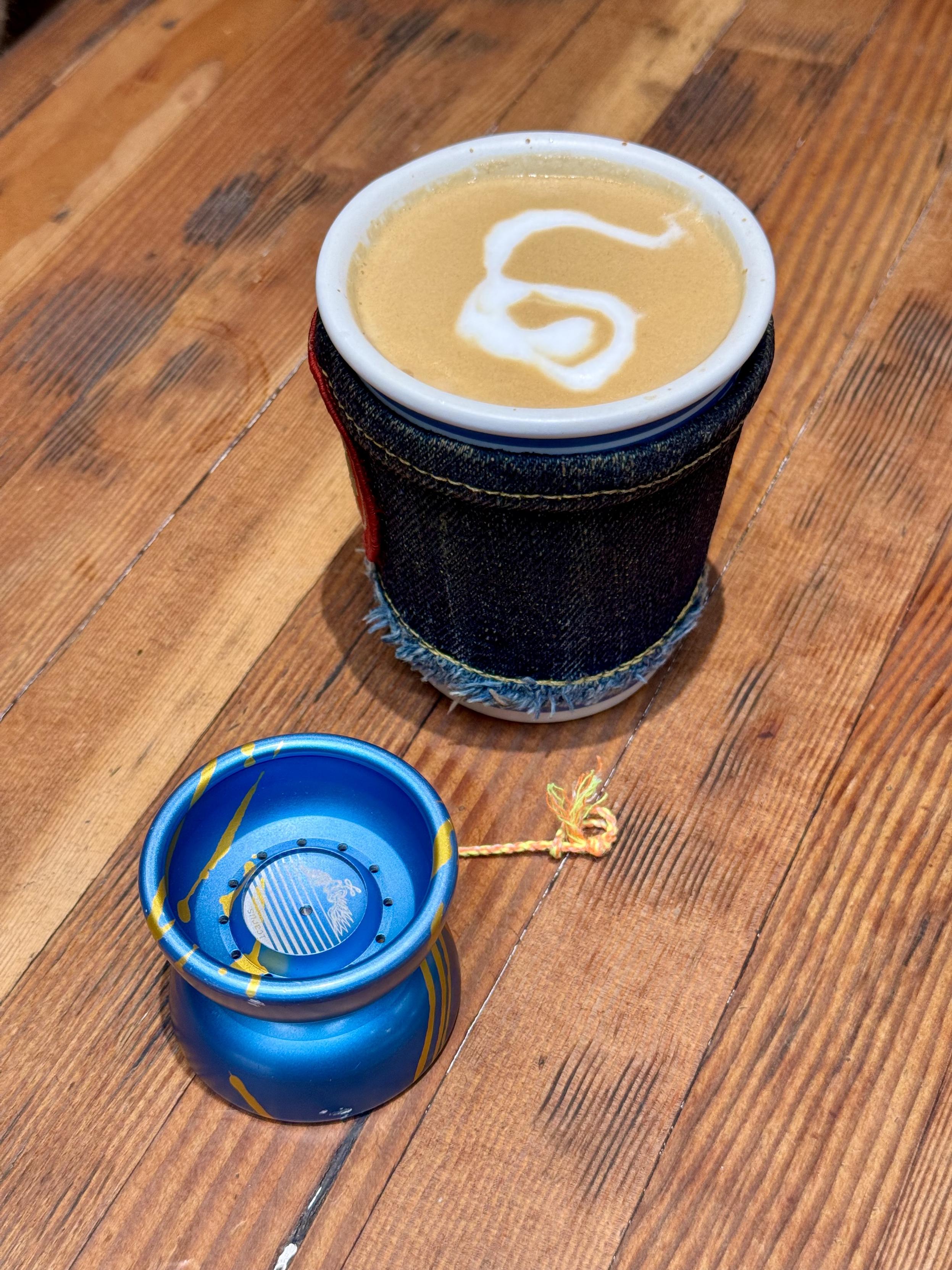 A yo-yo on a wooden table, sitting next to a cup of cappuccino. The barista who made this cappuccino got creative and made a yo-yo shaped piece of art in the foam. It's not perfect, but it's wobbly and cute.