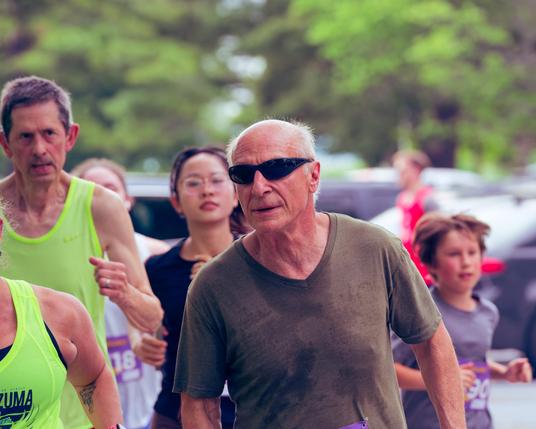 In the middle an older man with very short white hair and dark glasses and brown shirt has numerous runners in the background including men in high-viz tank tops, an asian woman and a male child