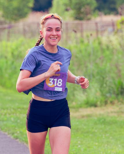 Woman with smile and hair braided with medium blue shirt, a little bit of belly showing and black shorts with different textured vegetation and a fence behind