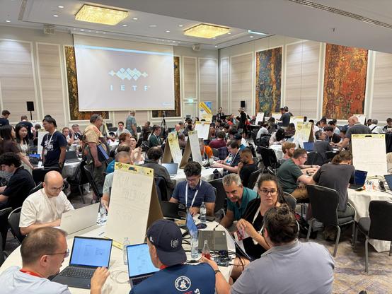 A busy conference room filled with attendees participating in discussions and collaborative work. There are multiple tables with laptops, papers, and signs featuring topics like XMPP and protocols. The IETF logo is visible on a screen in the back. 