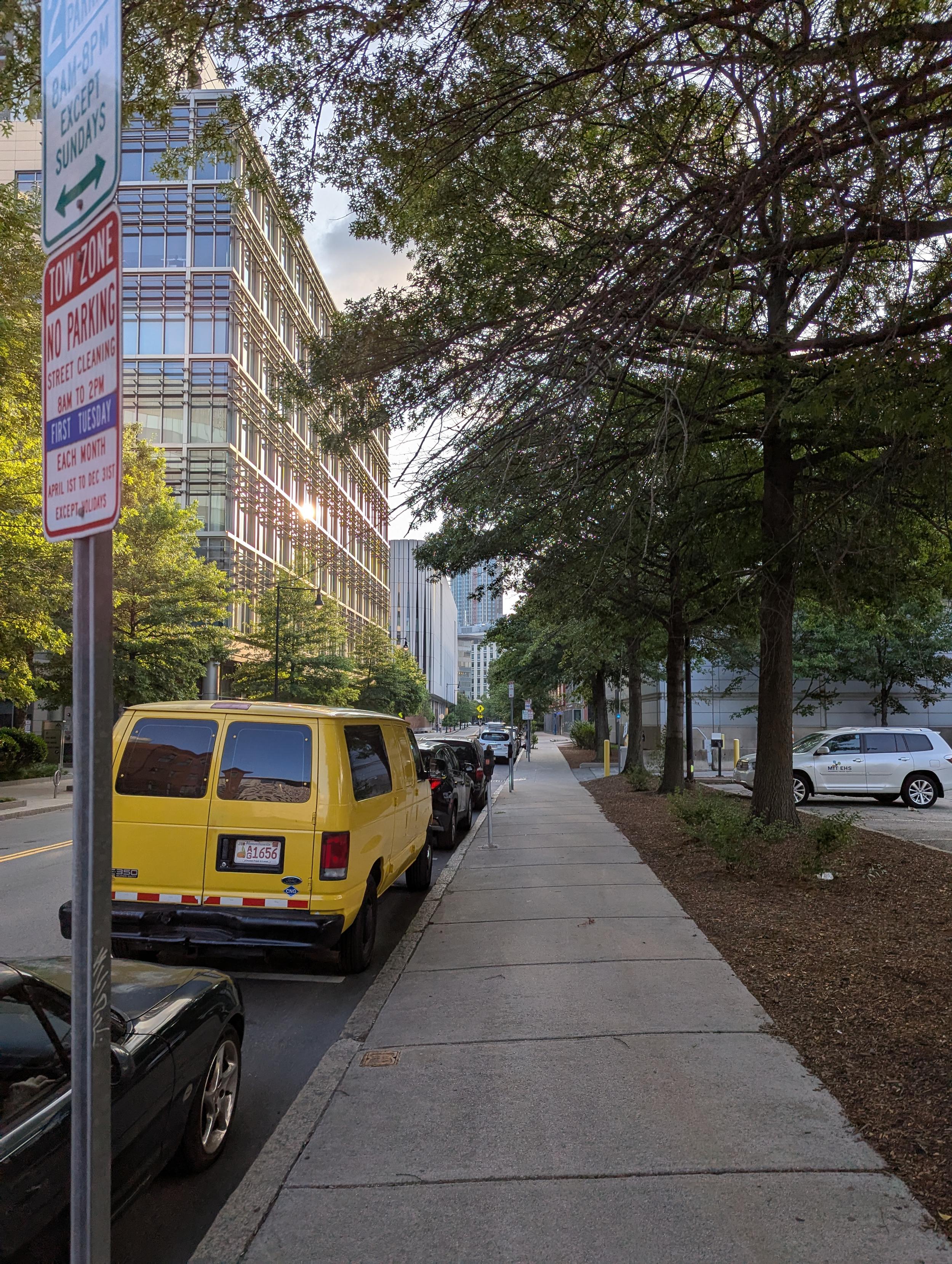 A photo of a quiet city street on a beautiful summer morning. Vehicles are lined up along the curb, and sunlight is reflecting off tall buildings but the street is still shaded.