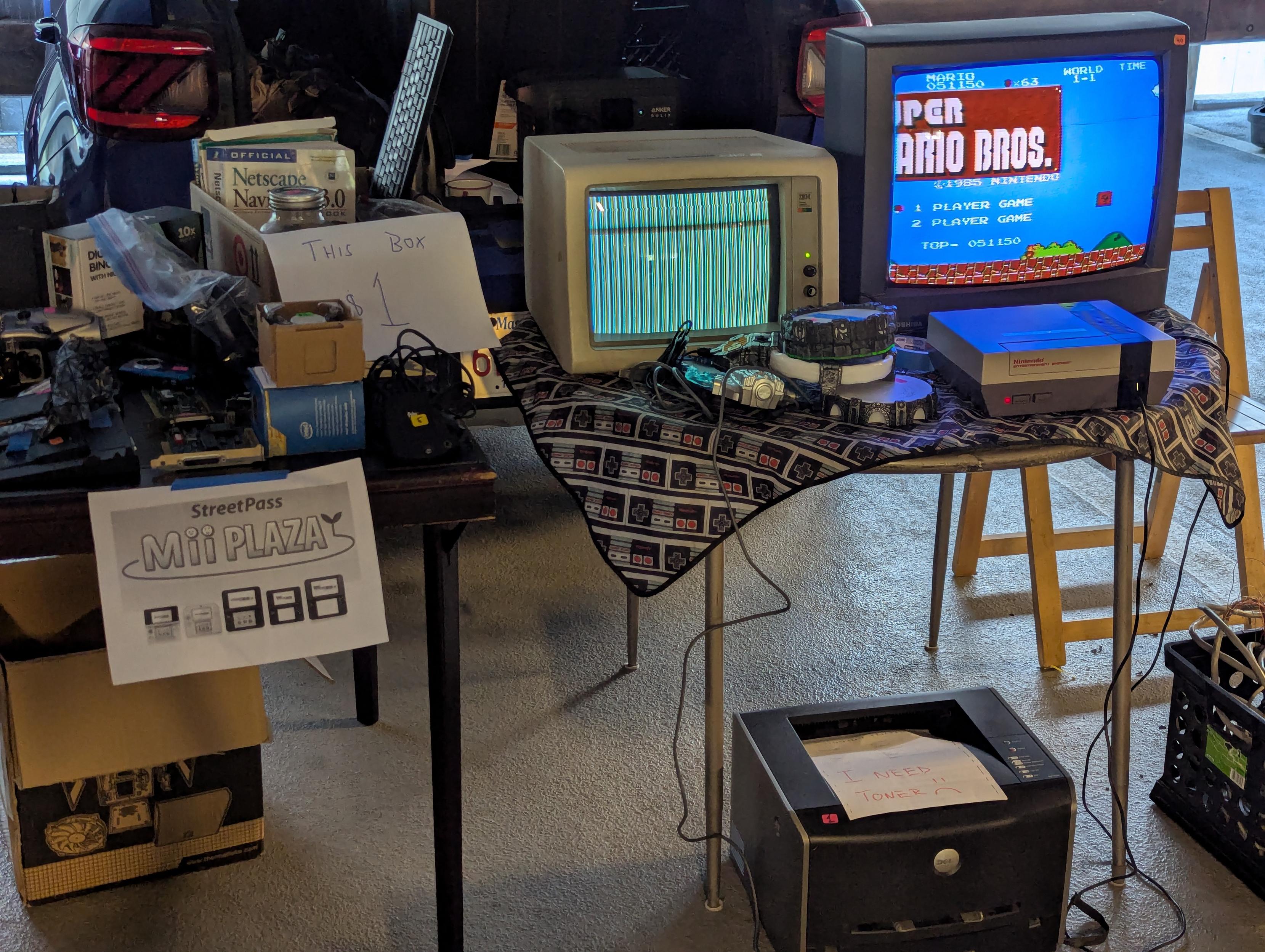 A photograph of a vendor's tables at the MIT Swapfest on 2026-07-19. The table on the left has an assortment of books, electronics, and cables. The table on the right has a CGA monitor displaying a test pattern, and a tube television showing Super Mario Bros. for the NES.