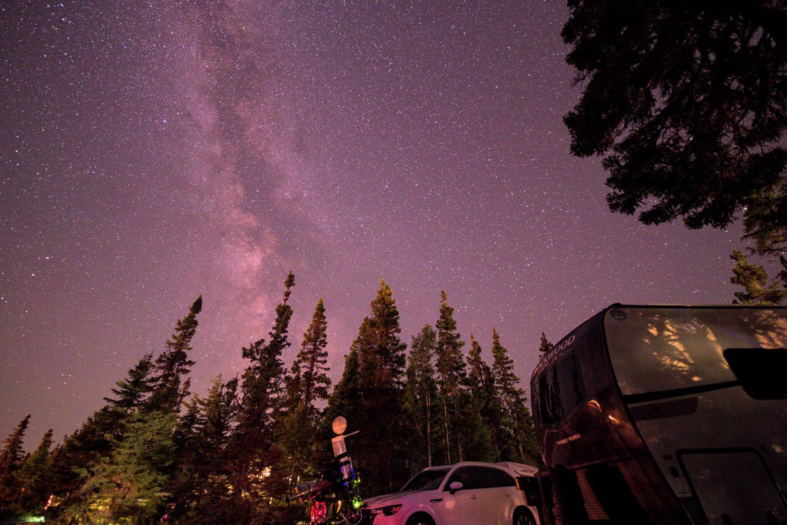 Night time image at a campground.  There are trees in the foreground. A parked car is located next to camper/caravan trailer on the right. A nearby tree is over hanging into the view at upper right. 

In near foreground is a telescope,  pointing up at the sky. 

Rising above the trees at center is the Milky Way. The dark lanes (dust obscuring starlight) of the Milky Way are evident. The Milky Way is brightest and slight orange-ish colour near the horizon. The rest of the sky is filled with many many small stars.

The sky has a slightly purple tinge because the photographers processing skills are suss 😆.