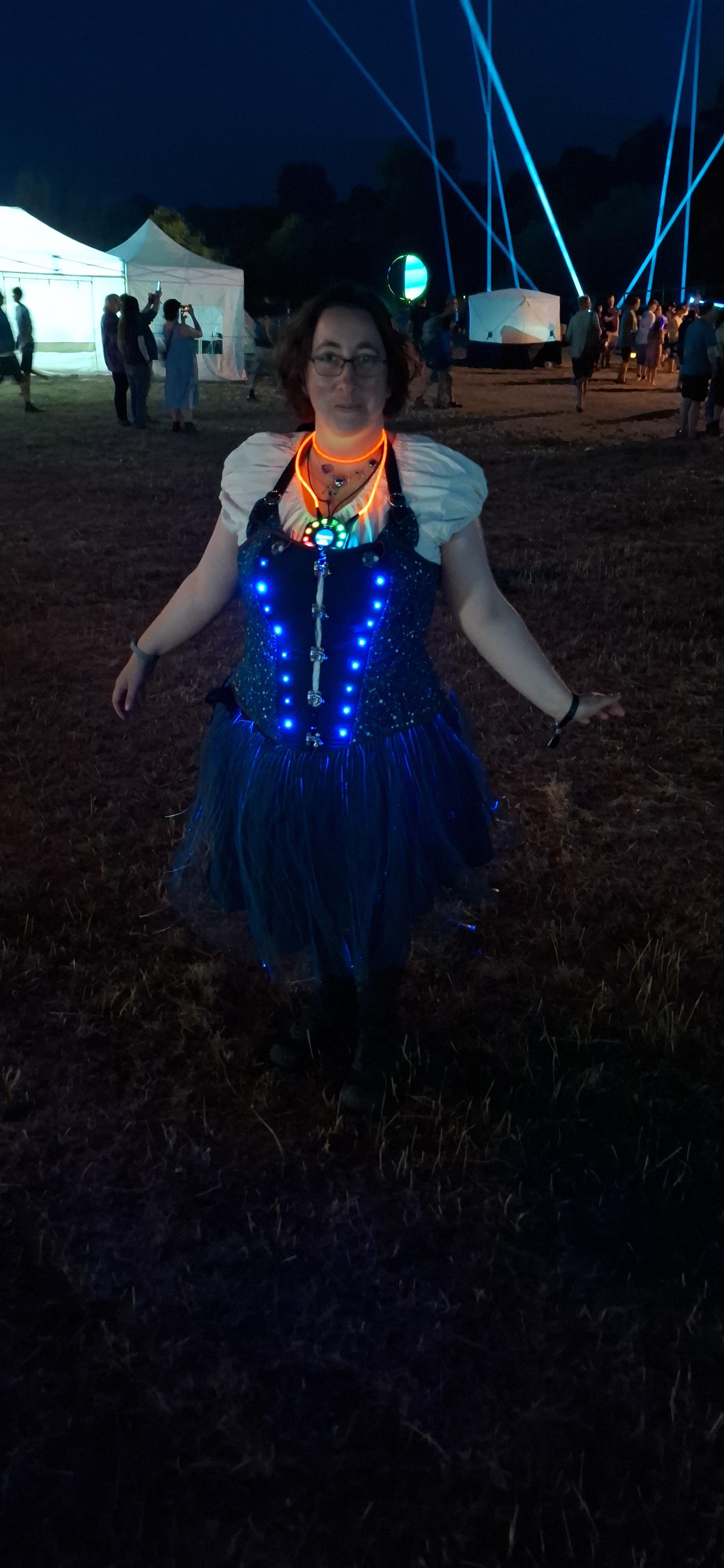 Charlie is wearing a corset with 18 blue LEDs along the sides of the front panel. The corset has  constellation patterns on the other fabric, abd the front panel is navy blue. They are also wearing a fibre optic skirt with blue light showimg faintly through some of it and a tildagon on a light up usb cable around their neck. They look very happy.