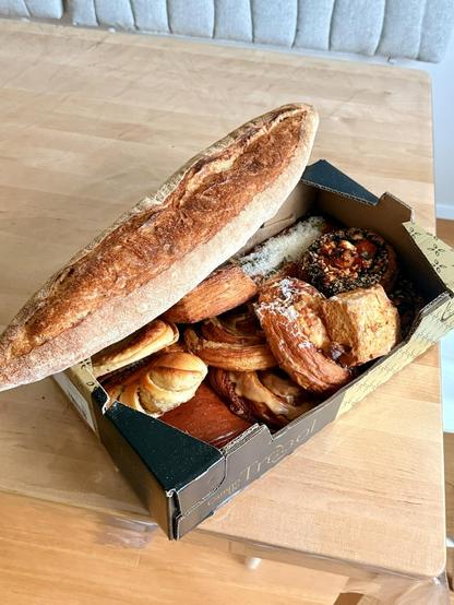 A box filled to the brim with delicious pastries and a giant loaf of French bread on top. 