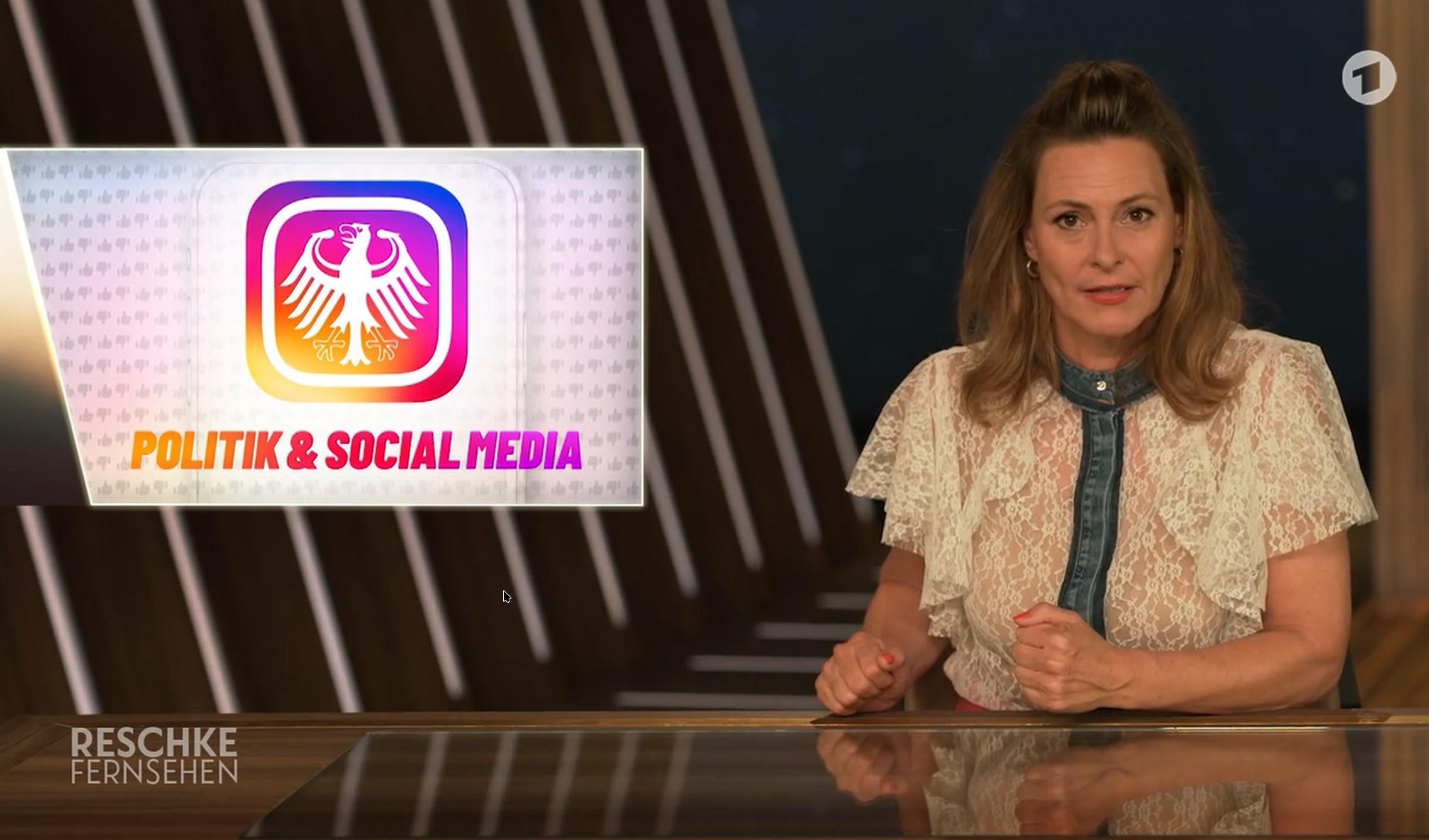 Ein Screenshot einer Fernsehsendung zeigt Anja Reschke hinter einem reflektierenden Schreibtisch und eine eingeblendete Grafik auf der linken Seite. Die Person (Anja Reschke)  mit langem braunem Haar trägt eine weiße Bluse mit Spitzenbesatz und blauem Kragen. In der Grafik ist ein weißes Adler-Symbol auf einem violett-orangen Farbverlauf zu sehen, darunter steht der Text „PO „POLITIK & SOCIAL MEDIA“. Am unteren linken Bildrand befindet sich der Text „RESCHKE FERNSEHEN“. Oben rechts ist zudem ein kreisförmiges Logo mit einer „1“ abgebildet.