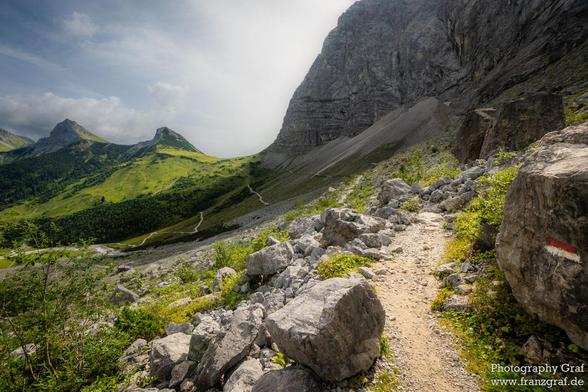 This image captures a breathtaking and rugged mountain landscape, evoking a sense of adventure and natural grandeur. A narrow, winding dirt trail cuts through the scene, leading the viewer's eye deeper into the majestic valley. The trail is flanked by rocky outcrops and patches of green vegetation, adding texture and contrast to the rugged terrain.
To the right of the trail, a steep, rocky cliff rises dramatically, its gray and brown hues showcasing the raw beauty of the mountain. The cliff's r…