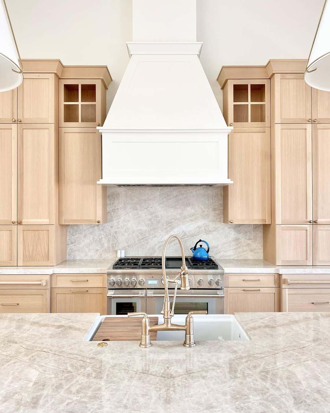 A beautiful white oak kitchen, showcasing a white vent hood over a stainless steel range. In the foreground is an island with a sink set into the stone countertop.
Look closely at how different pieces of the white oak are slightly different colors. They may be different due to using plywood panels combined with solid wood trim pieces, but it may also be due to the grain running either horizontally or vertically, depending on the piece.