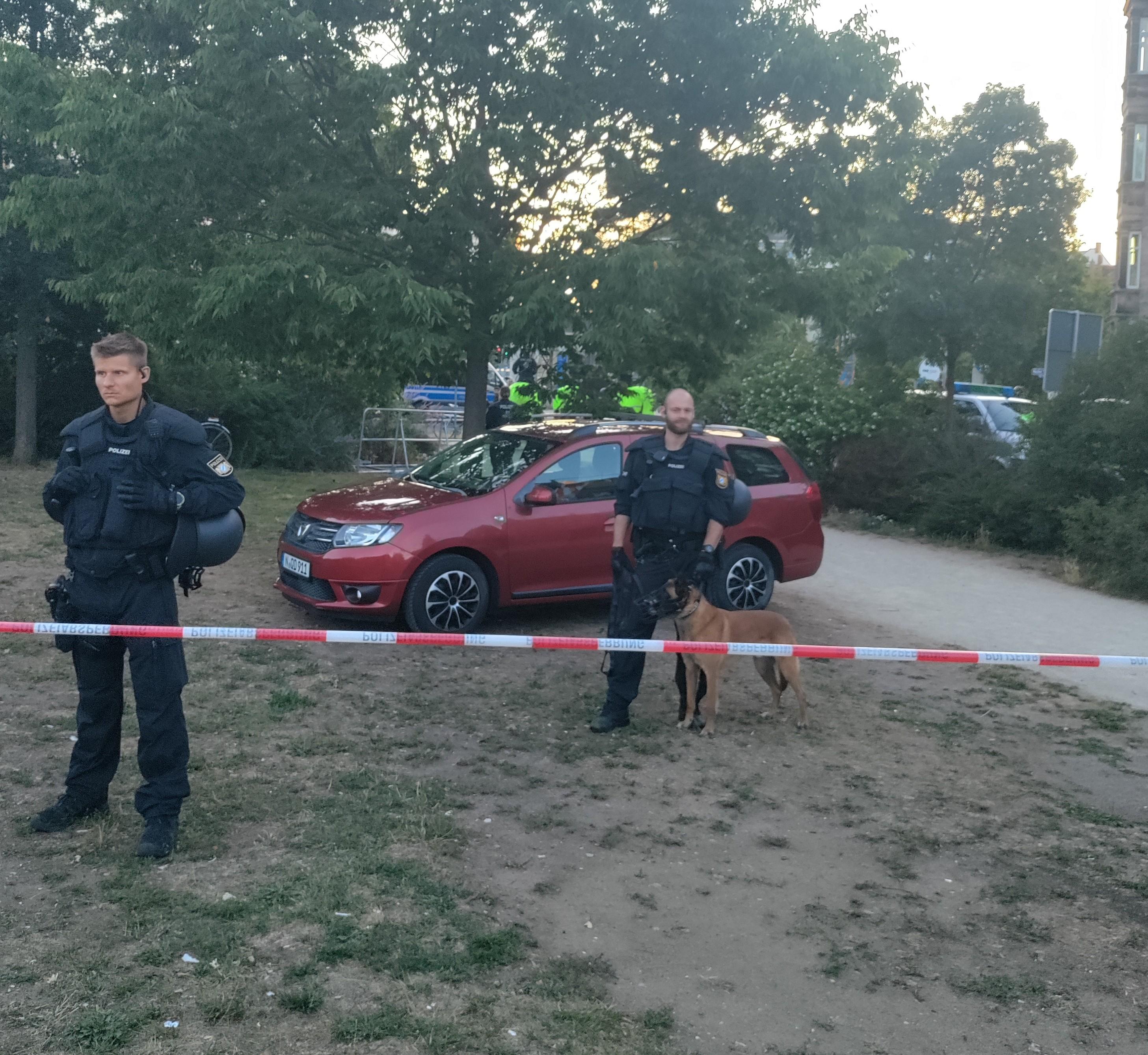 Die ganze Tierquälerei auf einem Blick; Das Auto von den Nazis, bewacht durch Polizist*innen mit Hund und Maulkorb, im Hintergrund auf der Straße hinter Bäumen sind mehrere gestressten Pferden zu sehen.
