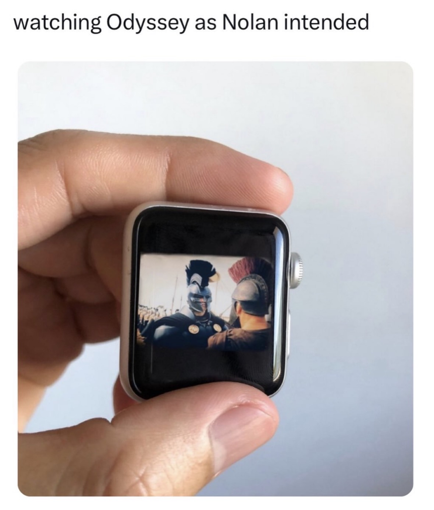 A hand holding a smartwatch displaying a scene from a historical movie where two soldiers in ancient Greek or Roman helmets are talking. The text above the image reads, "watching Odyssey as Nolan intended."