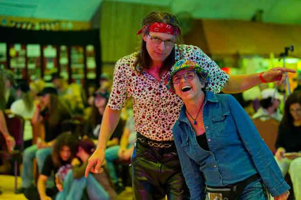 At the center a young man with rather long hair tied back in a bandanna and a white shirt with lots of red dots an dark pants,  in front on the right a laughing woman with a many colored hat and blue jacket with a crowd of spectators to a music event in back.